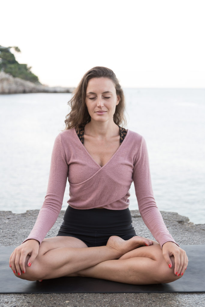 YOGA HINA posture de yoga en relaxation. Jambes en tailleur. Sur la plage. Haut rose. Zenitude.